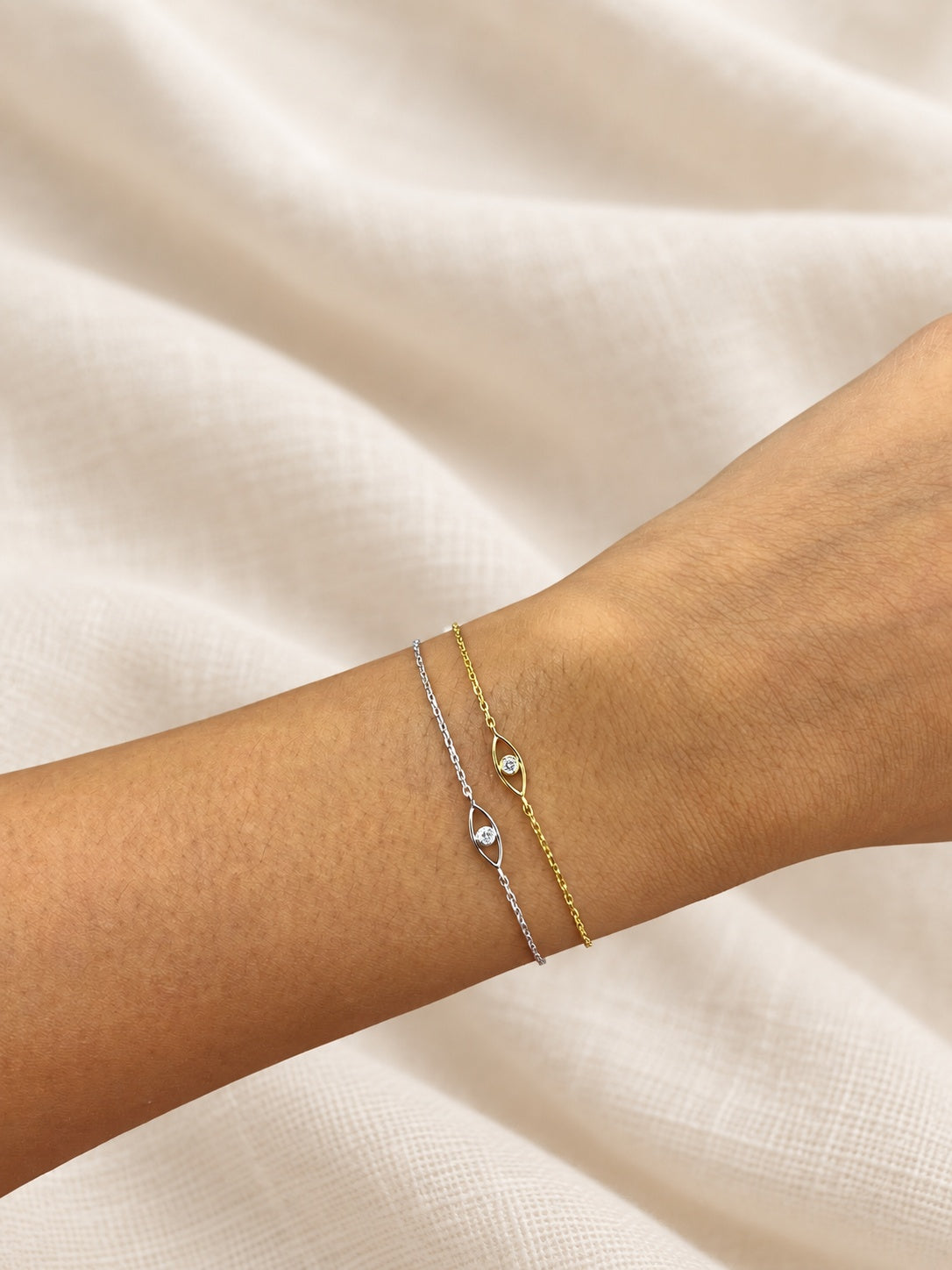 White-gold and yellow-gold evil-eye bracelets on a wrist against soft ivory linen