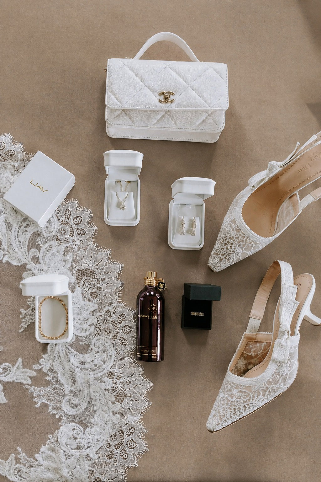LIAV jewellery in white presentation boxes beside lace bridal shoes and a white handbag on a Desert Rock taupe background